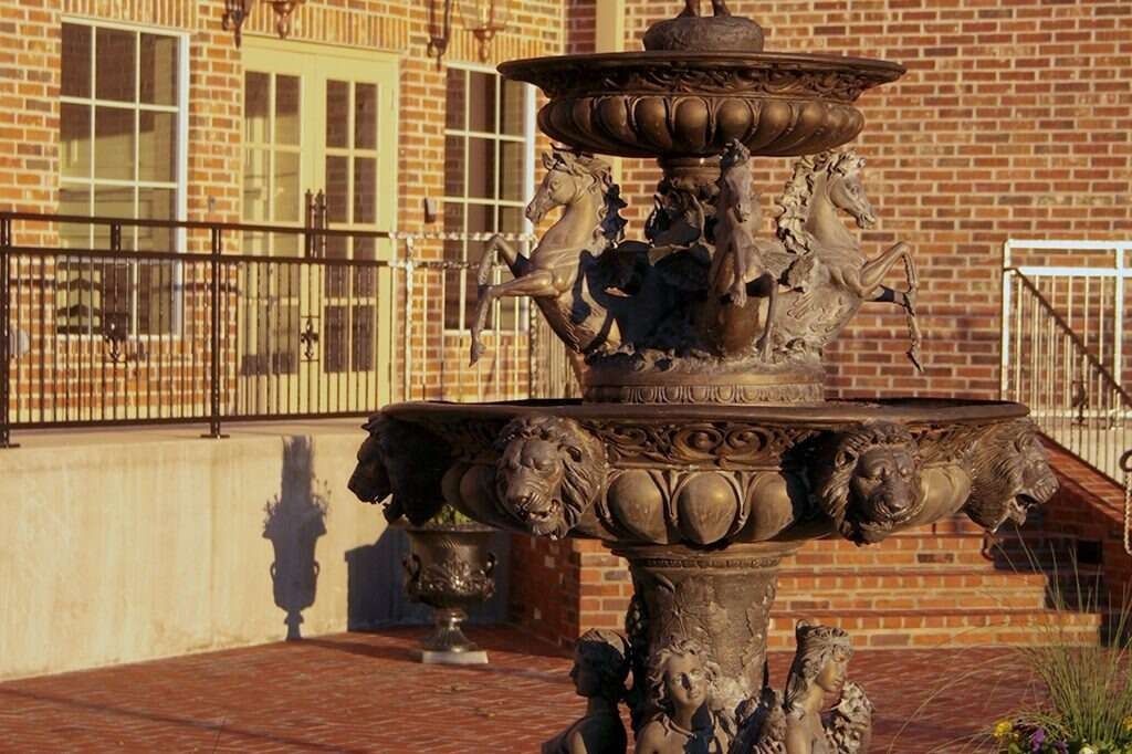 Ornate fountain on brick patio in front of a brick building, casting a long shadow.