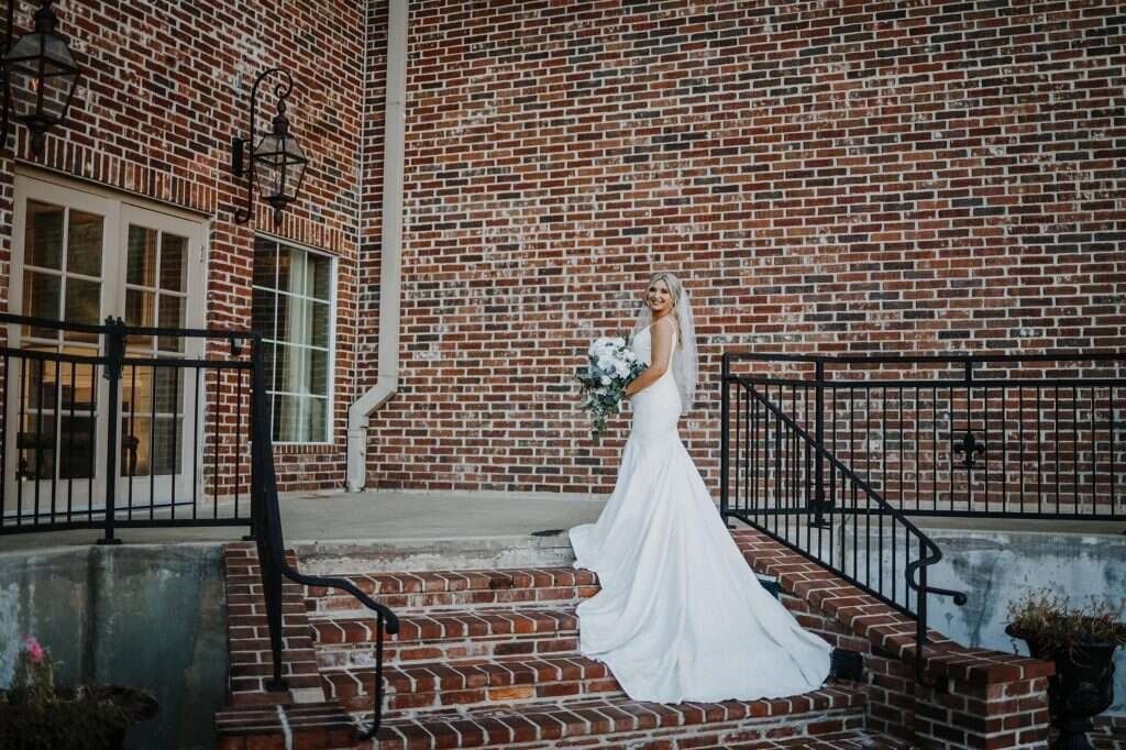 Bride in a white gown with a veil, holding flowers, standing on brick steps in front of a brick building.