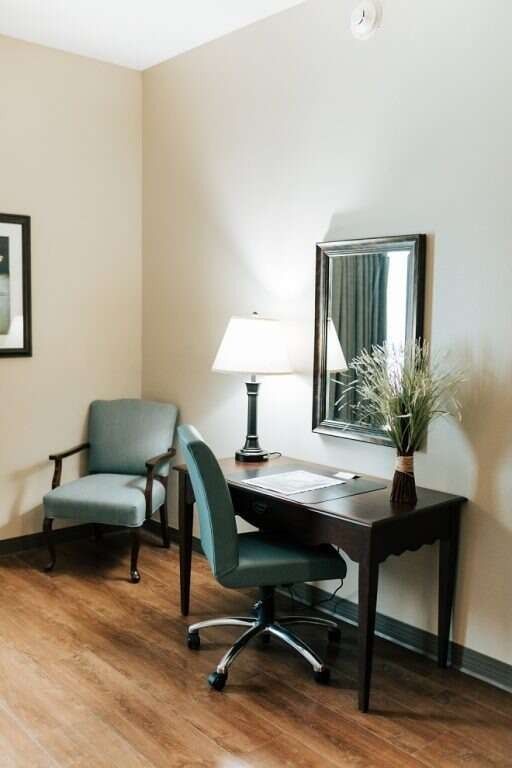 A desk with a chair, lamp, and mirror in a room with a blue chair, wooden floor and beige walls.