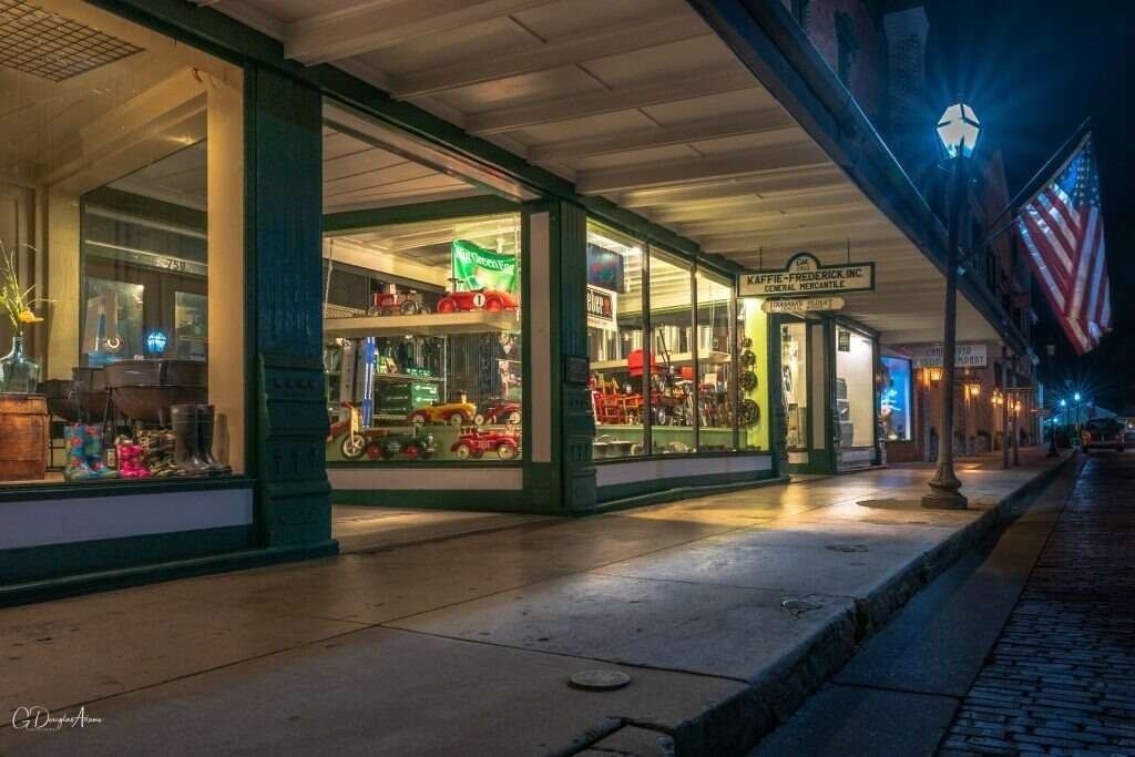 Nighttime view of shops with lit windows, American flag, and streetlights on cobblestone street.