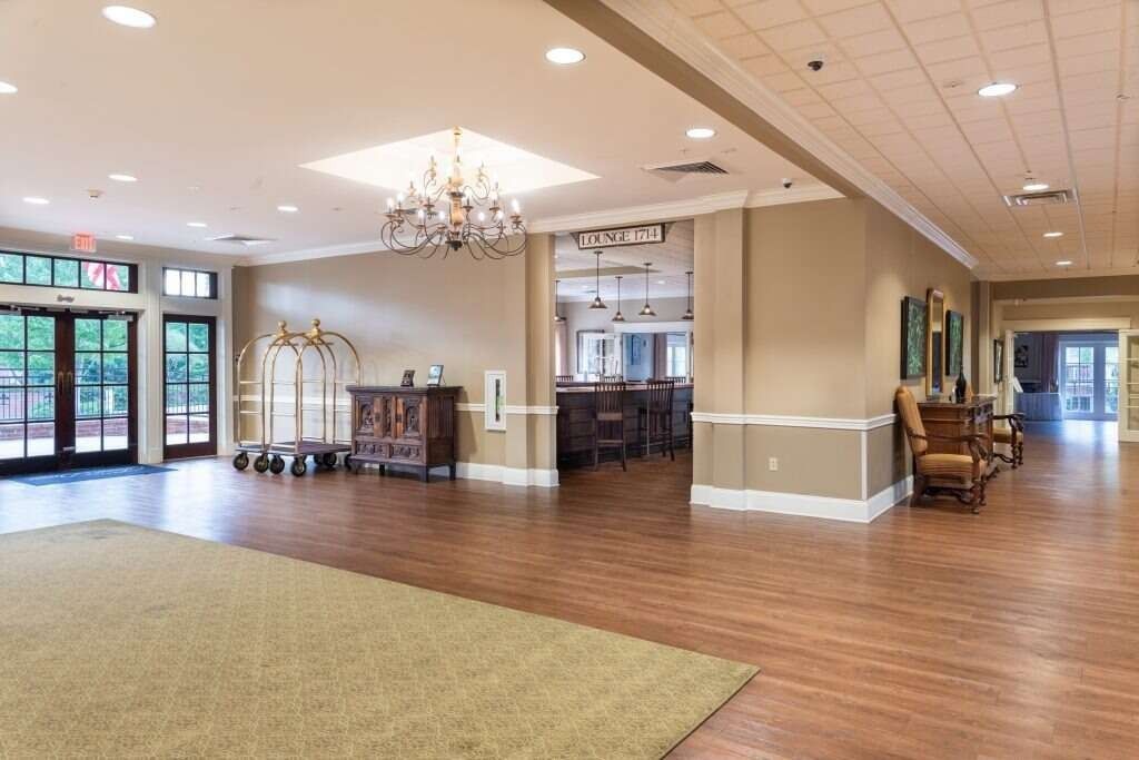 Large, well-lit interior lobby with wooden floors and a rug. Features a bar area, chandelier, and a hallway.