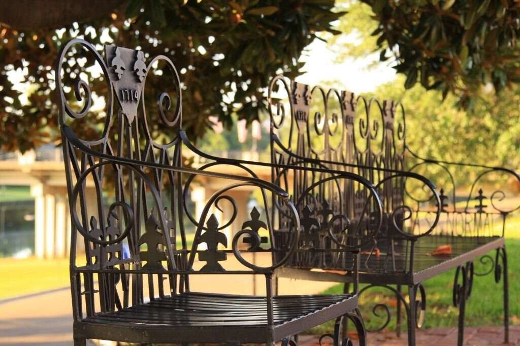 Ornate black wrought-iron bench in a park with a bridge visible in the background under a tree.