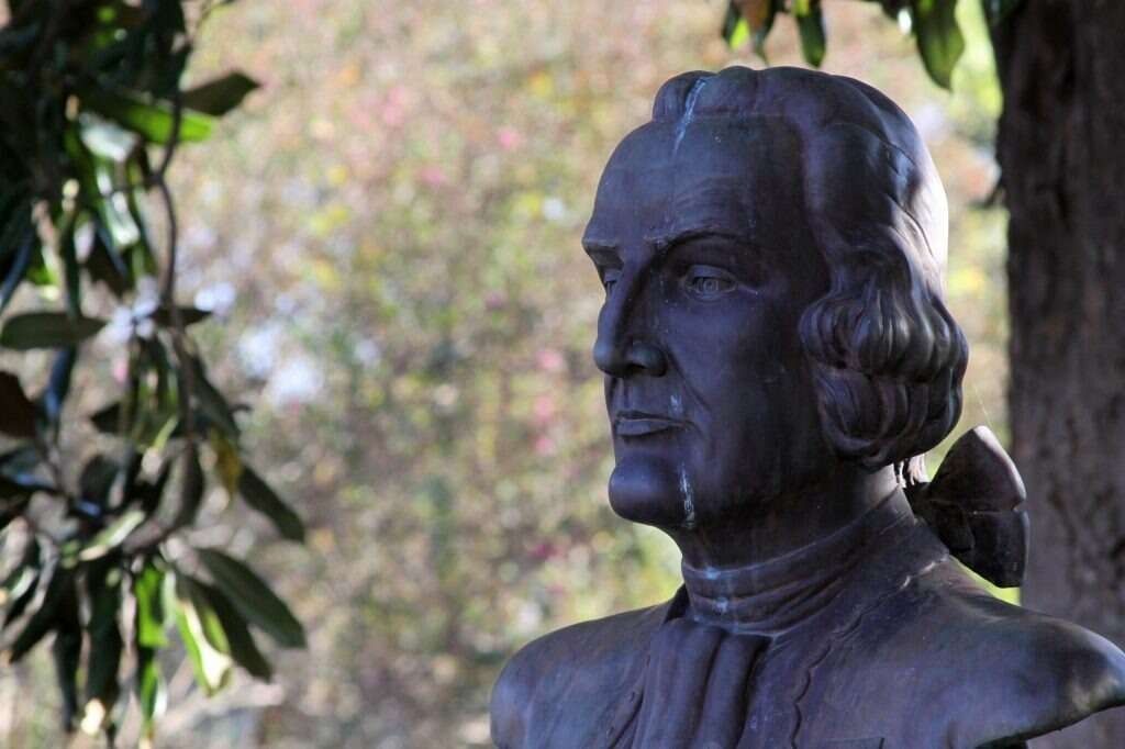 Bronze bust of a man outdoors, looking to the right. Blurred greenery in background.