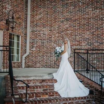 Bride in a white gown, veil, and holding a bouquet on brick steps of a building.